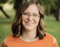 This picture is of a woman smiling, she has light brown hair and is wearing an orange t shirt. She is outside in nature, she is happy about her career in Tech.